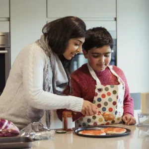 A woman and child prepare pizza in a kitchen. The woman guides the child in spreading sauce. The child wears a polka dot apron.